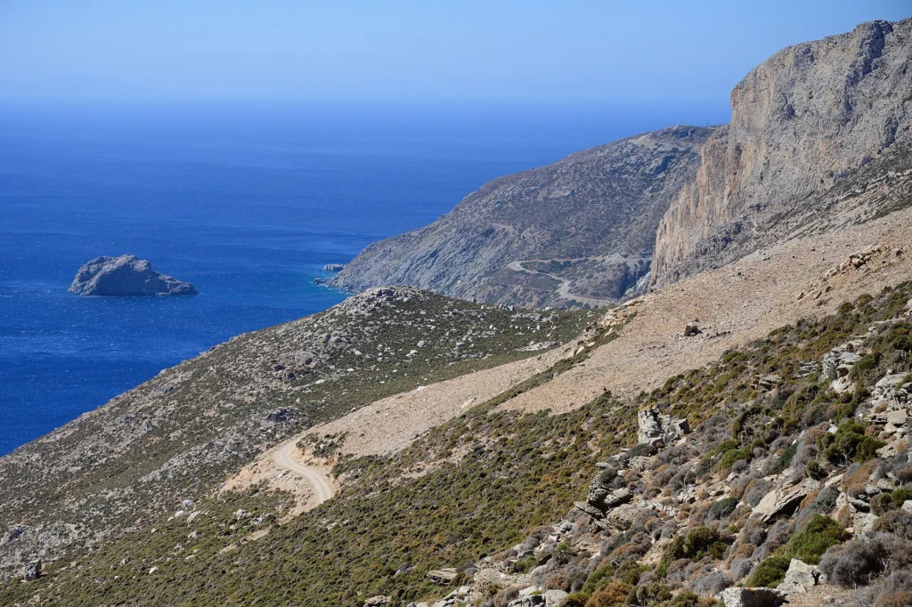Ξηρές πετρώδεις πλαγιές της Αμοργού πάνω από τη θάλασσα, με χωματόδρομο και βραχονησίδα στο βάθος