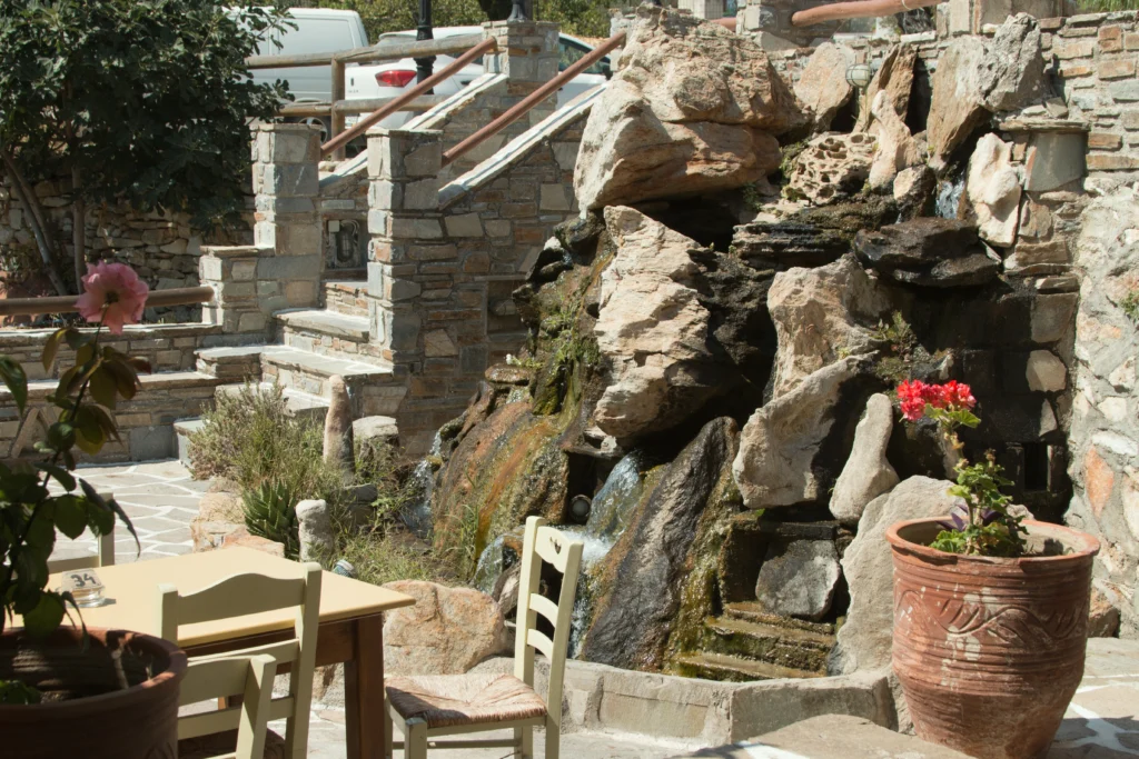 Τραπέζι με ψάθινες καρέκλες σε πλακόστρωτη αυλή ταβέρνας, δίπλα σε βράχια με νερό και γλάστρα με γεράνι