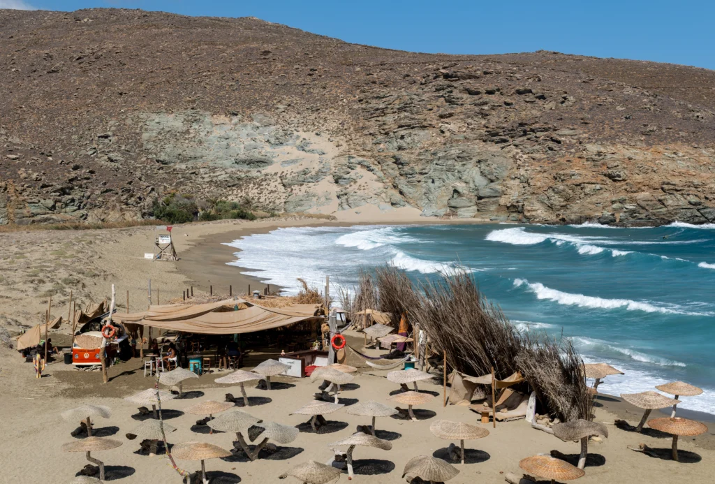 Beach bar με ψάθινες ομπρέλες σε αμμουδιά, με κύματα και ξερό λόφο στο βάθος, στη Μεγάλη Άμμο της Τήνου