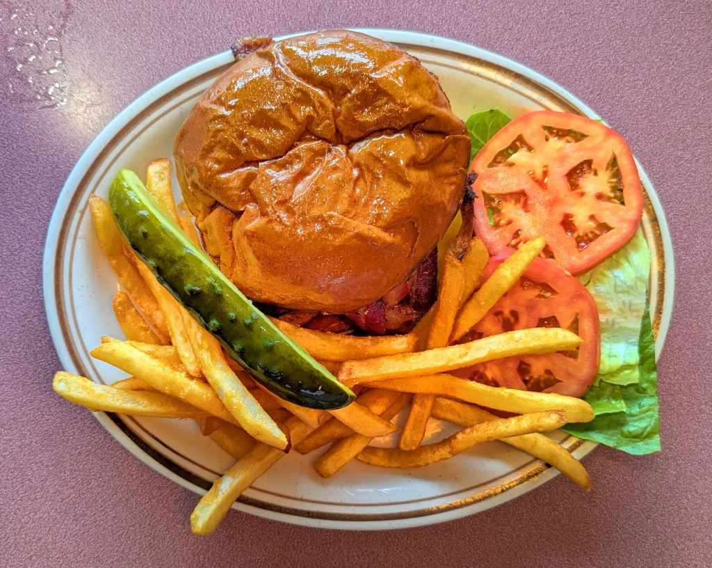 Burger με μπέικον σε γυαλιστερό ψωμάκι, με τηγανητές πατάτες, τουρσί, ντομάτα και μαρούλι σε οβάλ πιάτο