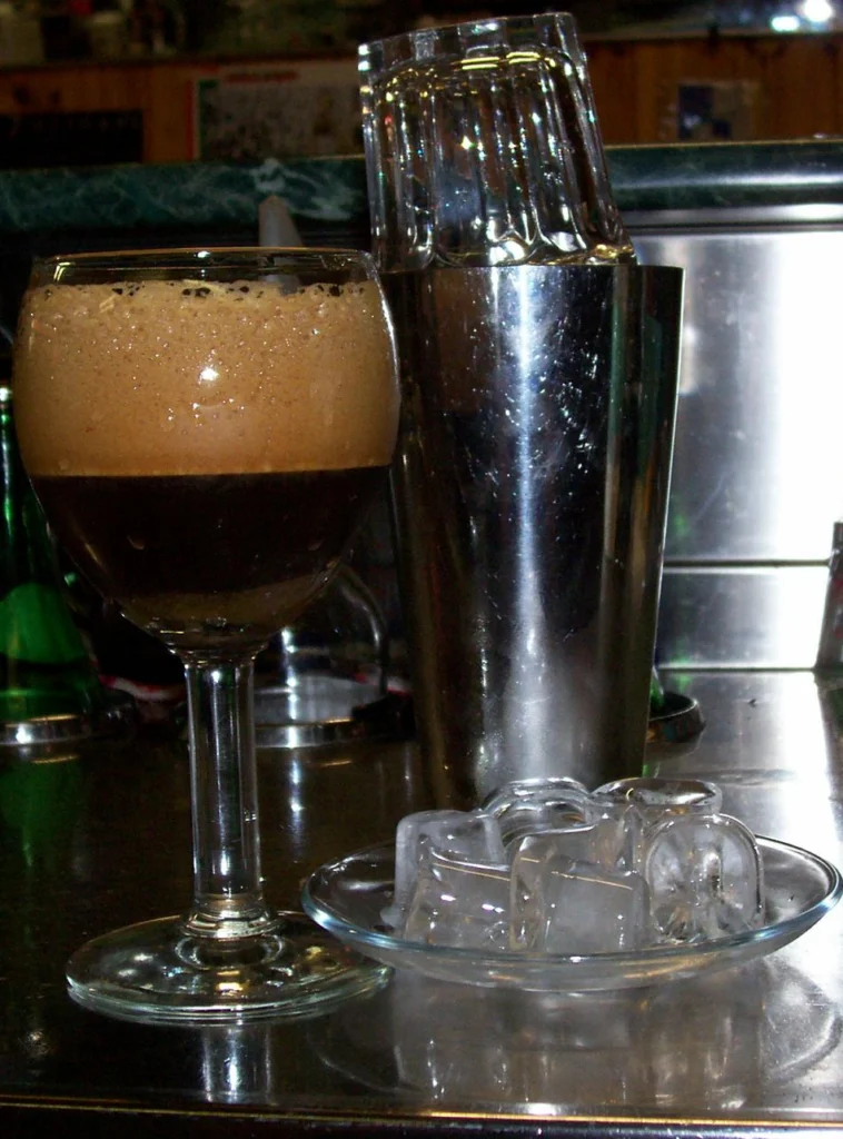 Ιταλικός caffè shakerato με αφρό σε ποτήρι με πόδι, δίπλα σε μεταλλικό σέικερ και παγάκια σε πιατάκι