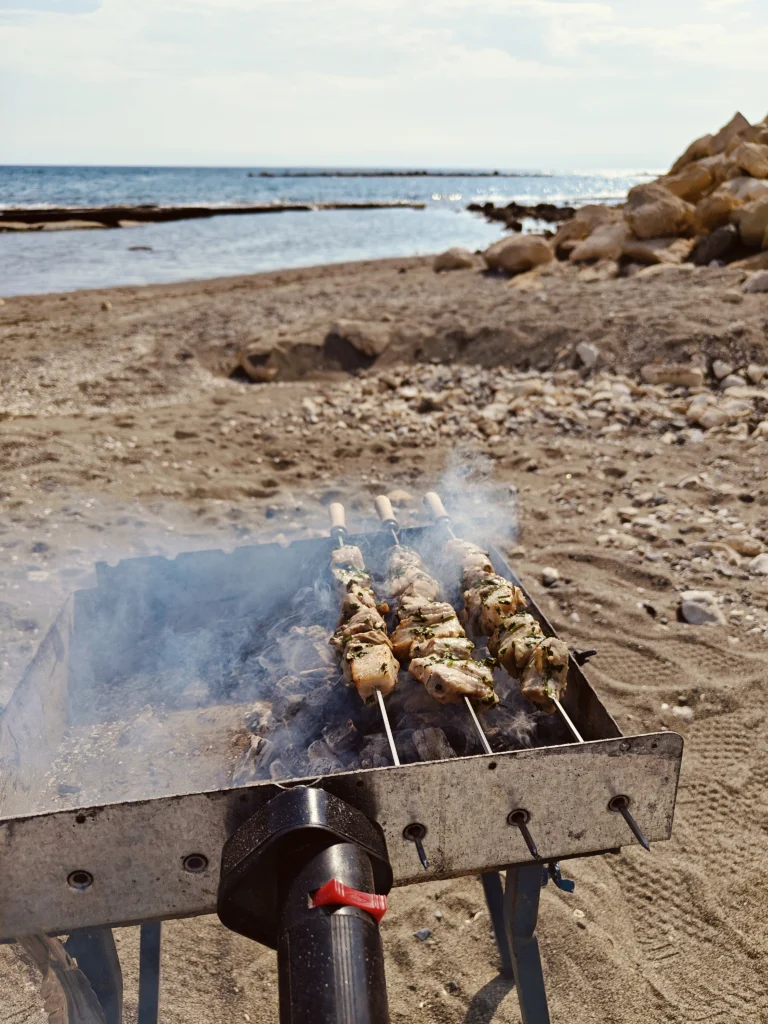 Τρία χοιρινά σουβλάκια με μυρωδικά ψήνονται σε μεταλλική ψησταριά με κάρβουνα, με τη θάλασσα στο βάθος