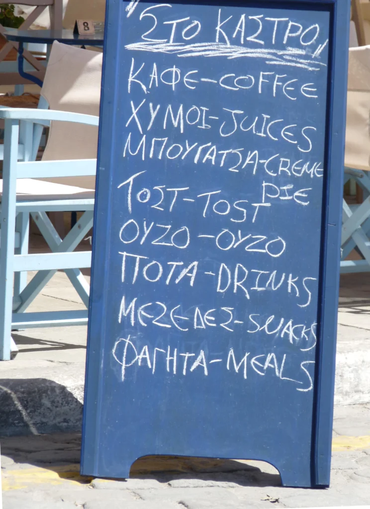 Μπλε μαυροπίνακας με είδη σε ελληνικά και αγγλικά: καφές, χυμοί, μπουγάτσα, ούζο, μεζέδες, φαγητά