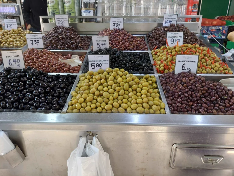 Πάγκος με δέκα είδη πράσινες και μαύρες ελιές σε μεταλλικές λεκάνες με ταμπελάκια τιμών