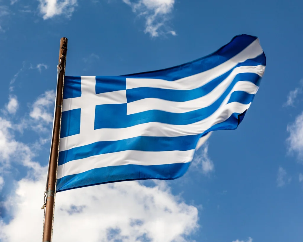 Η ελληνική σημαία κυματίζει σε μεταλλικό ιστό κάτω από γαλάζιο ουρανό με λευκά σύννεφα