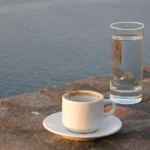 Φλιτζάνι ελληνικού καφέ σε πιατάκι και ποτήρι νερό πάνω σε πέτρα, με τη θάλασσα πίσω