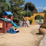 Αυλή ταβέρνας με παιδική χαρά: παιδικό σπιτάκι, τσουλήθρα, κούνιες και πλαστικό τραπεζάκι με πάγκους