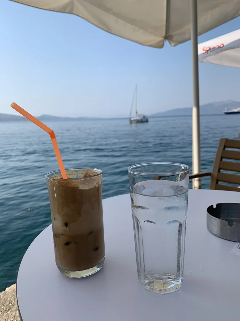 Φραπέ σε ψηλό ποτήρι με καλαμάκι και ποτήρι νερό πάνω σε στρογγυλό τραπέζι δίπλα στη θάλασσα