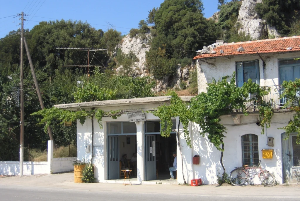 Ασβεστωμένο κτίριο καφενείου χωριού με κληματαριά, ξύλινες πόρτες και ποδήλατο, με βουνό στο βάθος