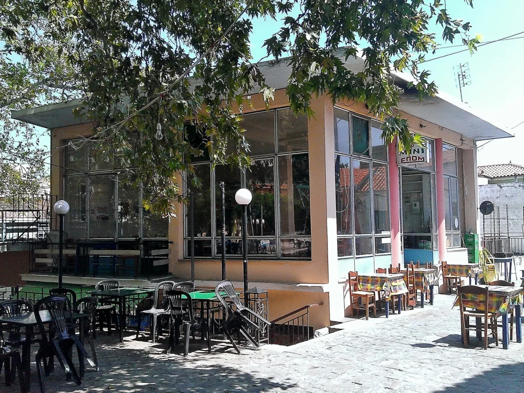 Καφετέρια με τραπεζάκια και καρέκλες στην αυλή κάτω από δέντρα σε χωριό της Λέσβου