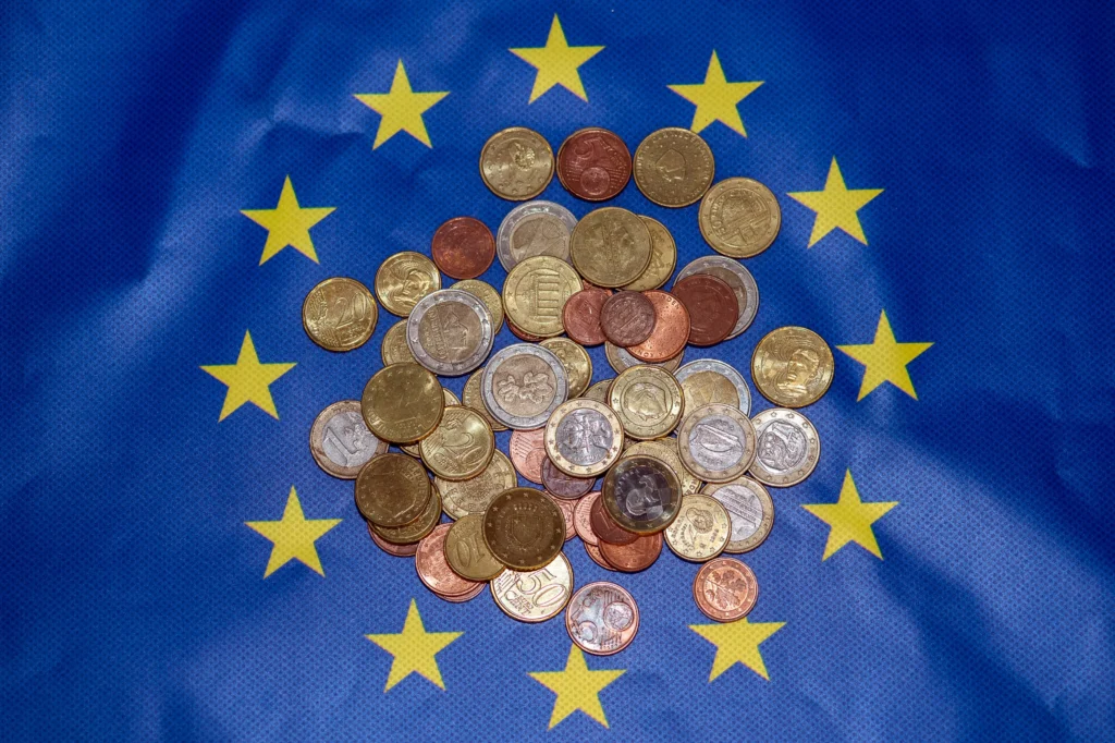 Κέρματα ευρώ διαφόρων αξιών μαζεμένα στο κέντρο μιας μπλε σημαίας της ΕΕ με κίτρινα αστέρια