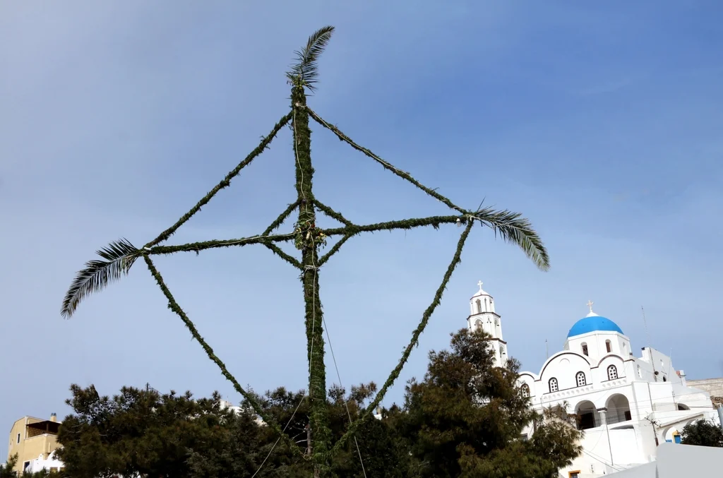 Ψηλός σταυρός στολισμένος με κλαδιά σε πλατεία της Σαντορίνης, με λευκή εκκλησία με μπλε τρούλο πίσω