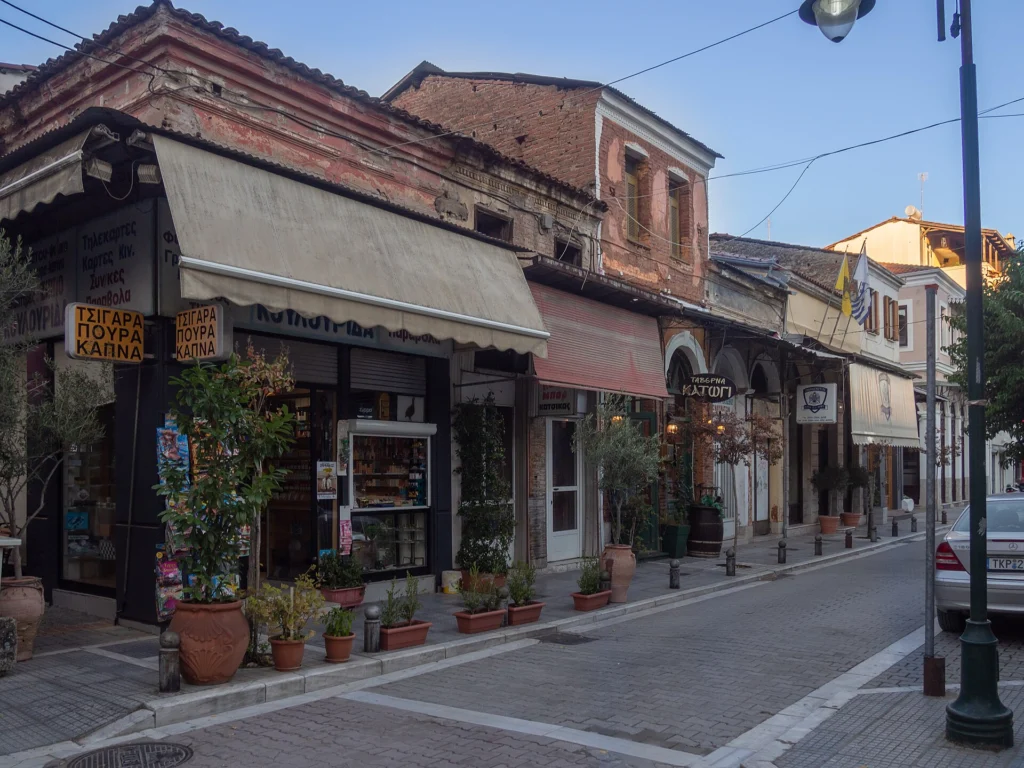 Παλιά διώροφα κτίρια με τέντες και ταμπέλες ταβέρνας σε λιθόστρωτο δρόμο της συνοικίας Μανάβικα στα Τρίκαλα