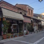 Παλιά διώροφα κτίρια με τέντες και ταμπέλες ταβέρνας σε λιθόστρωτο δρόμο της συνοικίας Μανάβικα στα Τρίκαλα