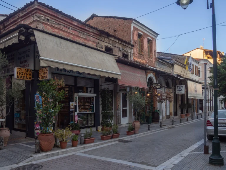 Παλιά διώροφα κτίρια με τέντες και ταμπέλες ταβέρνας σε λιθόστρωτο δρόμο της συνοικίας Μανάβικα στα Τρίκαλα