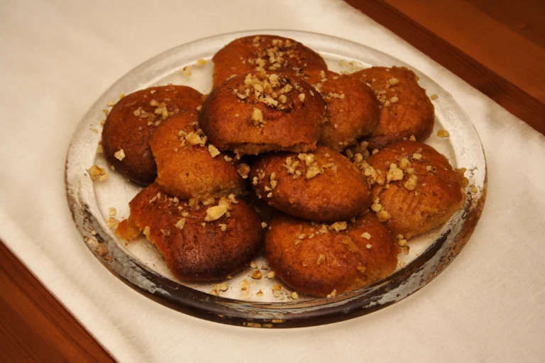 Γυάλινη πιατέλα με μελομακάρονα σιροπιασμένα και πασπαλισμένα με τριμμένο καρύδι πάνω σε λευκό τραπεζομάντιλο
