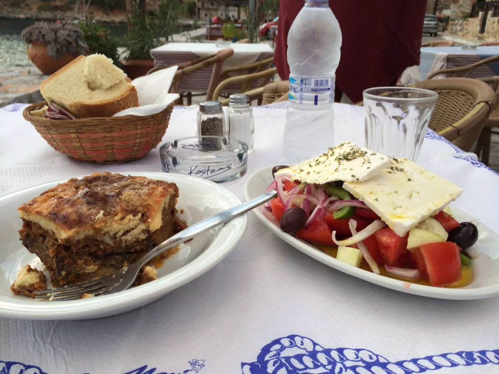 Μουσακάς και χωριάτικη σαλάτα με φέτα σε τραπέζι ταβέρνας, με ψωμί και ποτήρια νερού