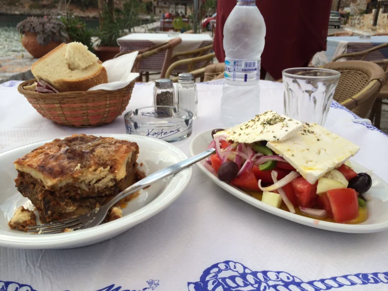 Μουσακάς και χωριάτικη σαλάτα με φέτα σε τραπέζι ταβέρνας, με ψωμί και ποτήρια νερού