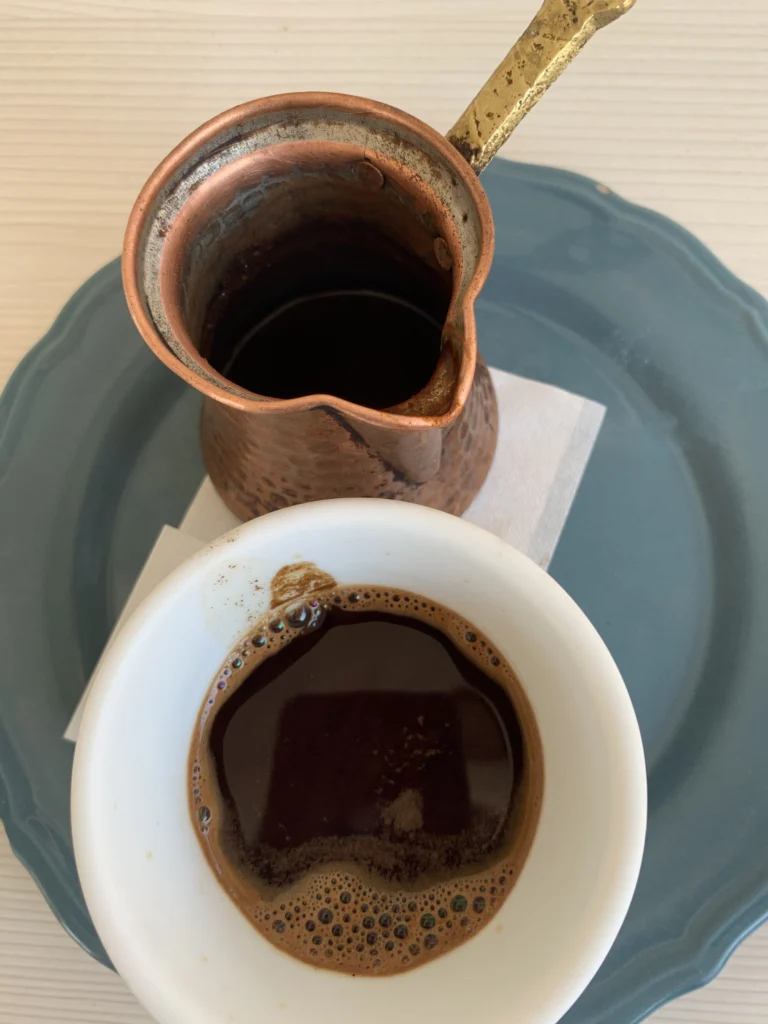 Χάλκινο μπρίκι με υπολείμματα καφέ δίπλα σε φλιτζάνι ελληνικού με αφρό, πάνω σε μπλε πιάτο