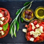 Κριθαρένιο παξιμάδι με ψιλοκομμένη ντομάτα και κύβους φέτας, δίπλα σε φέτες ντομάτας, ελιές και μπολ με ελαιόλαδο