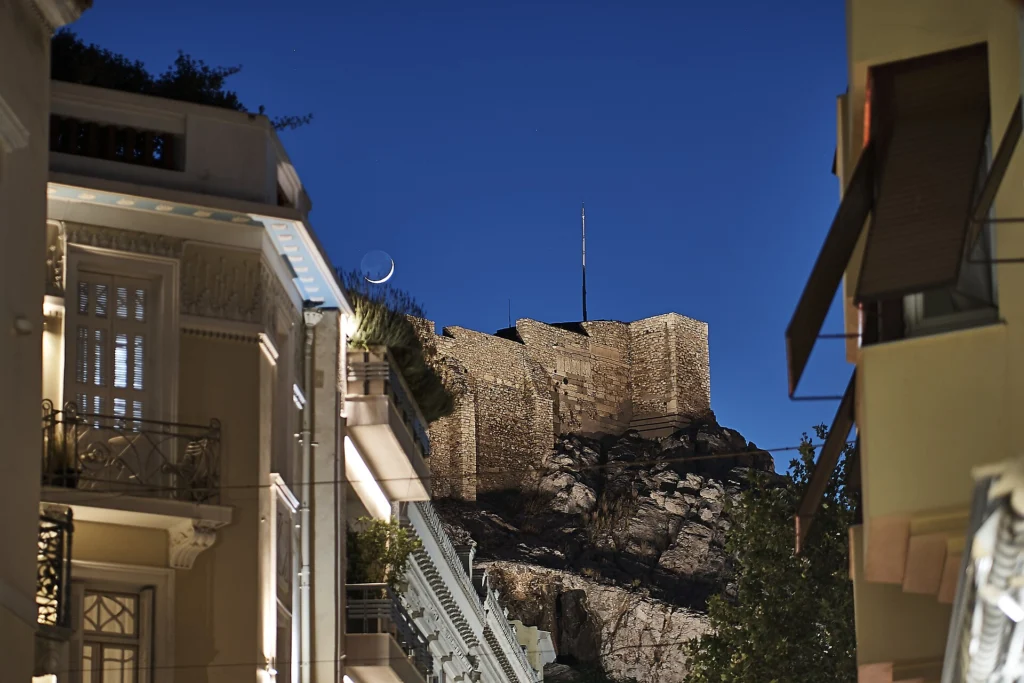 Νυχτερινή άποψη της Πλάκας με τα τείχη της Ακρόπολης και μισοφέγγαρο πάνω από τα σπίτια