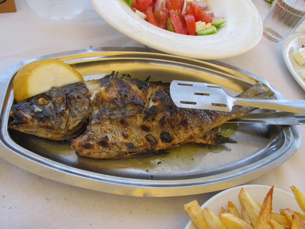Ολόκληρο ψητό ψάρι με φέτα λεμονιού σε μεταλλική πιατέλα, με χωριάτικη σαλάτα και τηγανητές πατάτες