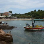Μικρό ελληνικό ψαροκάικο με κίτρινα δίχτυα δεμένο σε ήσυχο κόλπο, με παραλία και σπίτια στο βάθος