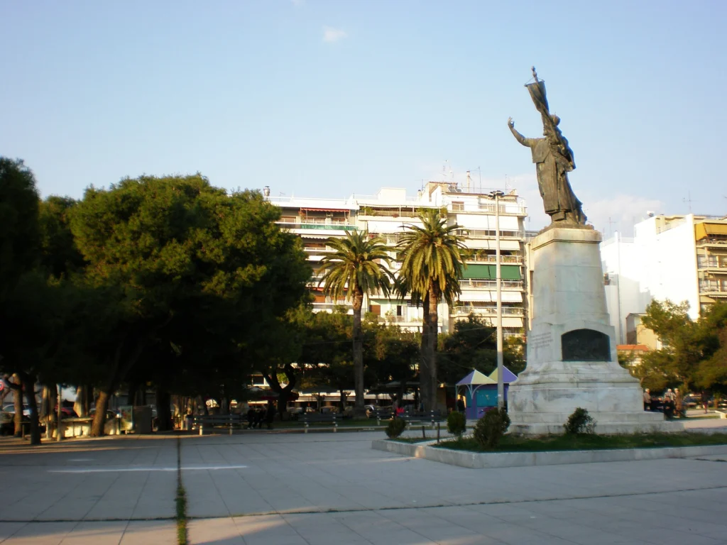 Η πλατεία Υψηλών Αλωνίων στην Πάτρα με φοίνικες, δέντρα, πολυκατοικίες και το άγαλμα του Παλαιών Πατρών Γερμανού