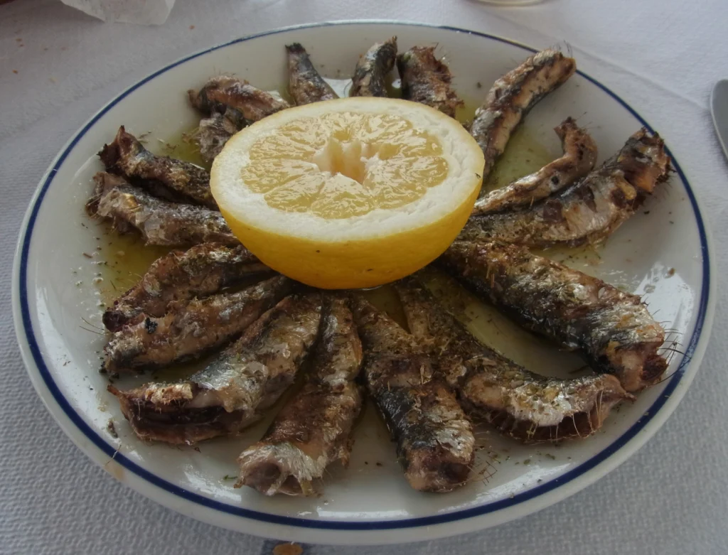 Ψητές σαρδέλες σε κύκλο γύρω από μισό λεμόνι, με λάδι και ρίγανη, σε λευκό πιάτο με μπλε ρίγα