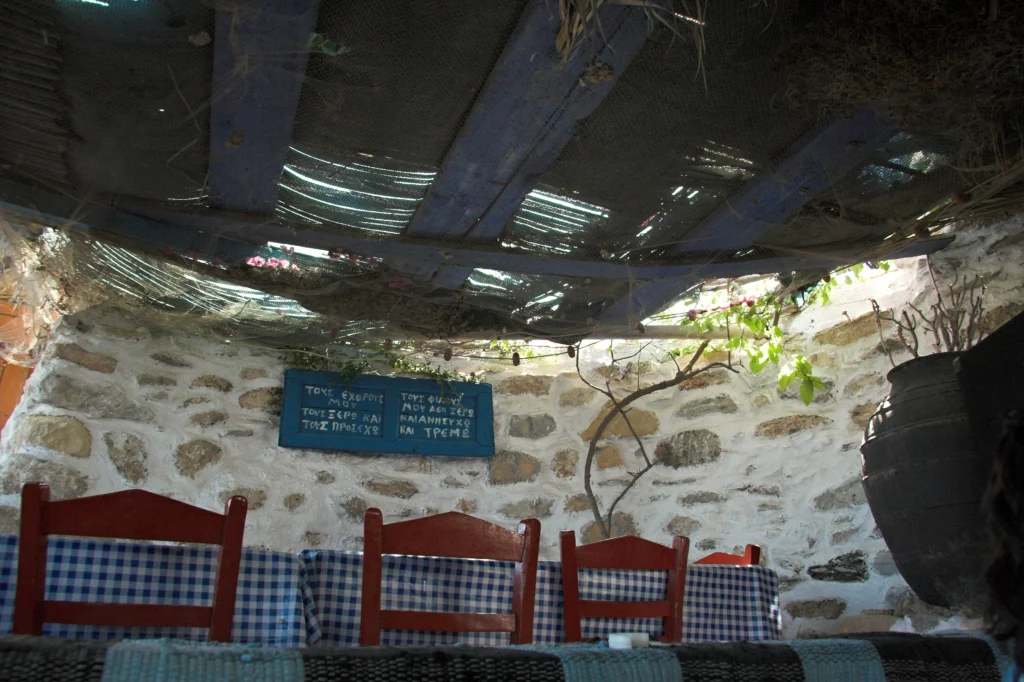 Εσωτερικό ταβέρνας με καρό τραπεζομάντιλα, κόκκινες καρέκλες και πέτρινο τοίχο με επιγραφή