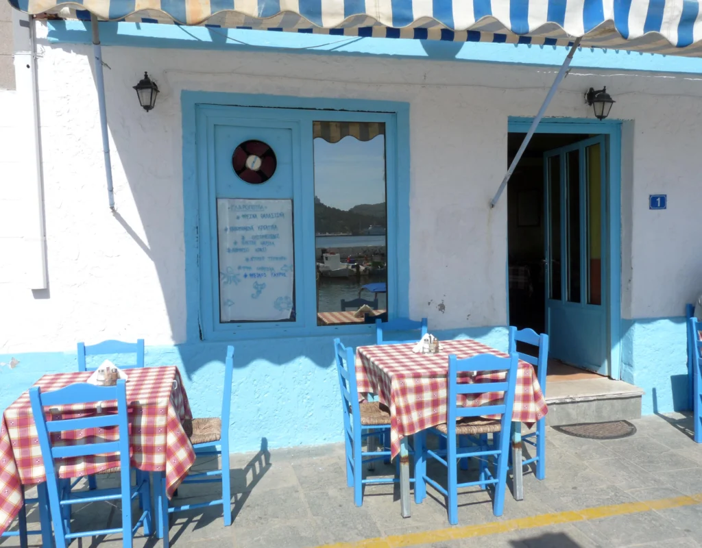 Πρόσοψη ταβέρνας με μπλε κουφώματα, χειρόγραφη λίστα πιάτων στο τζάμι και τραπέζια με καρό τραπεζομάντιλα