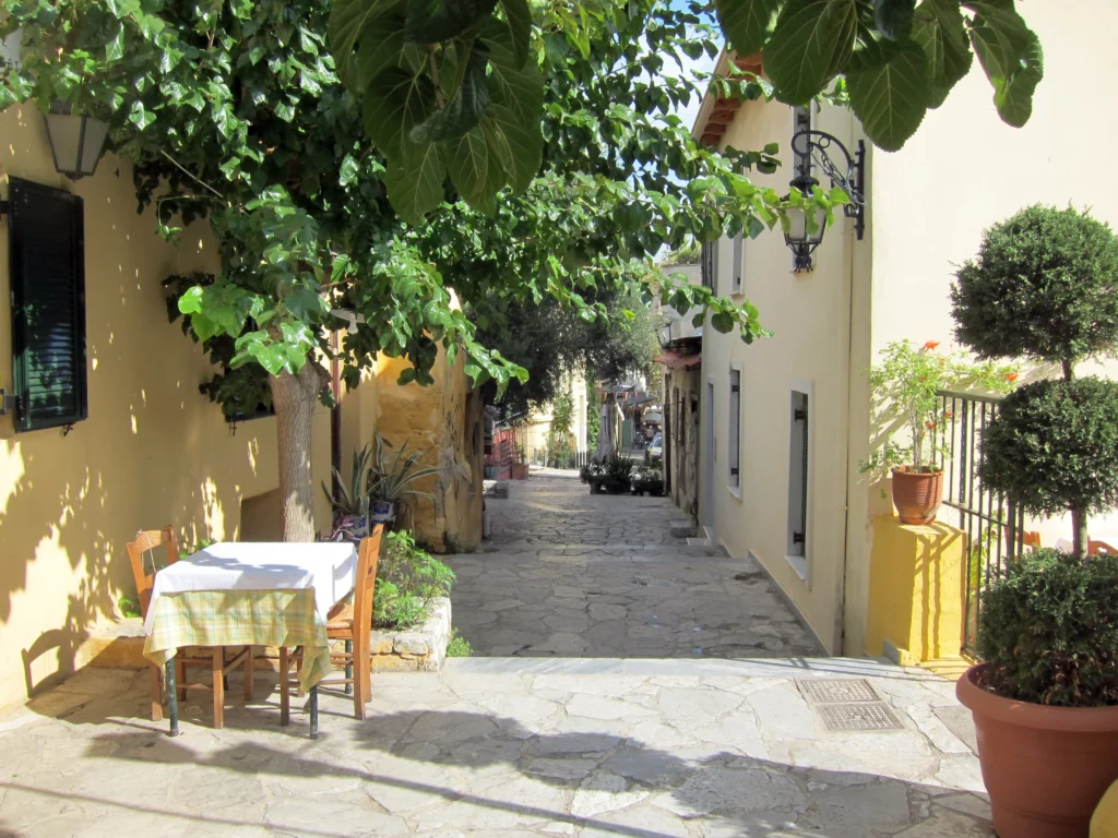 Άδεια τραπέζια ταβέρνας με τραπεζομάντιλα σε πλακόστρωτο σοκάκι της Πλάκας με γλάστρες και δέντρα