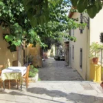 Άδεια τραπέζια ταβέρνας με τραπεζομάντιλα σε πλακόστρωτο σοκάκι της Πλάκας με γλάστρες και δέντρα