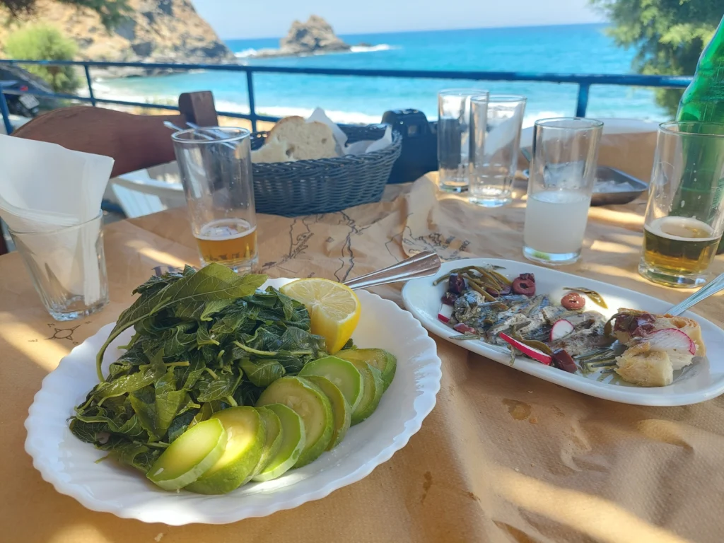 Τραπέζι ταβέρνας με βραστά χόρτα, κολοκυθάκια, θαλασσινό μεζέ, ούζο και μπίρα, με θέα τη θάλασσα στην Ικαρία