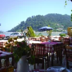 Τραπέζια ταβέρνας με καρό τραπεζομάντιλα και ομπρέλα, με θάλασσα και πράσινο λόφο στο βάθος, στη Θάσο