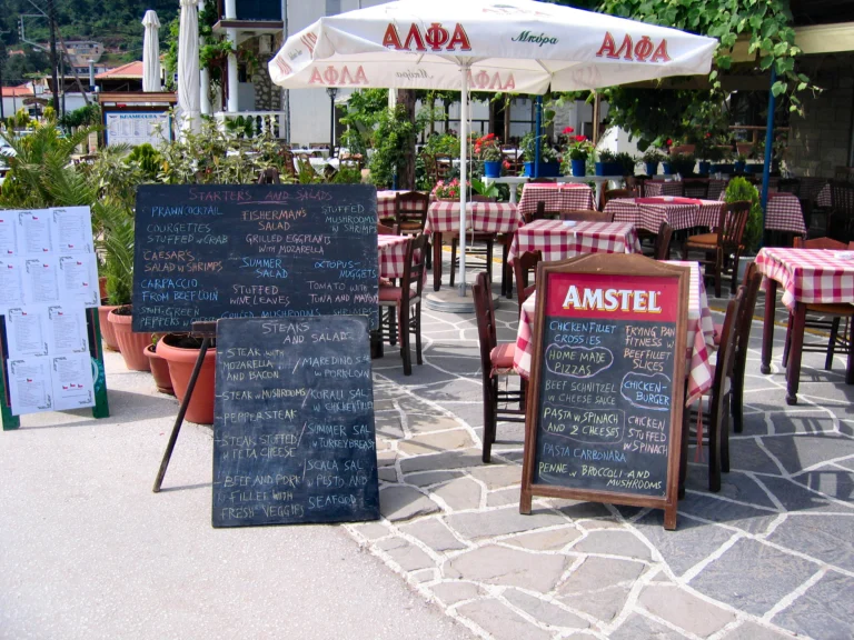 Πίνακες με κιμωλία και αναρτημένοι κατάλογοι στην είσοδο ταβέρνας στη Θάσο, με τραπέζια με καρό τραπεζομάντιλα