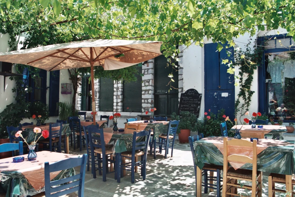 Φωτογραφία αυλής ταβέρνας στη Νάξο με μπλε καρέκλες, στρωμένα τραπέζια, ομπρέλα και κληματαριά