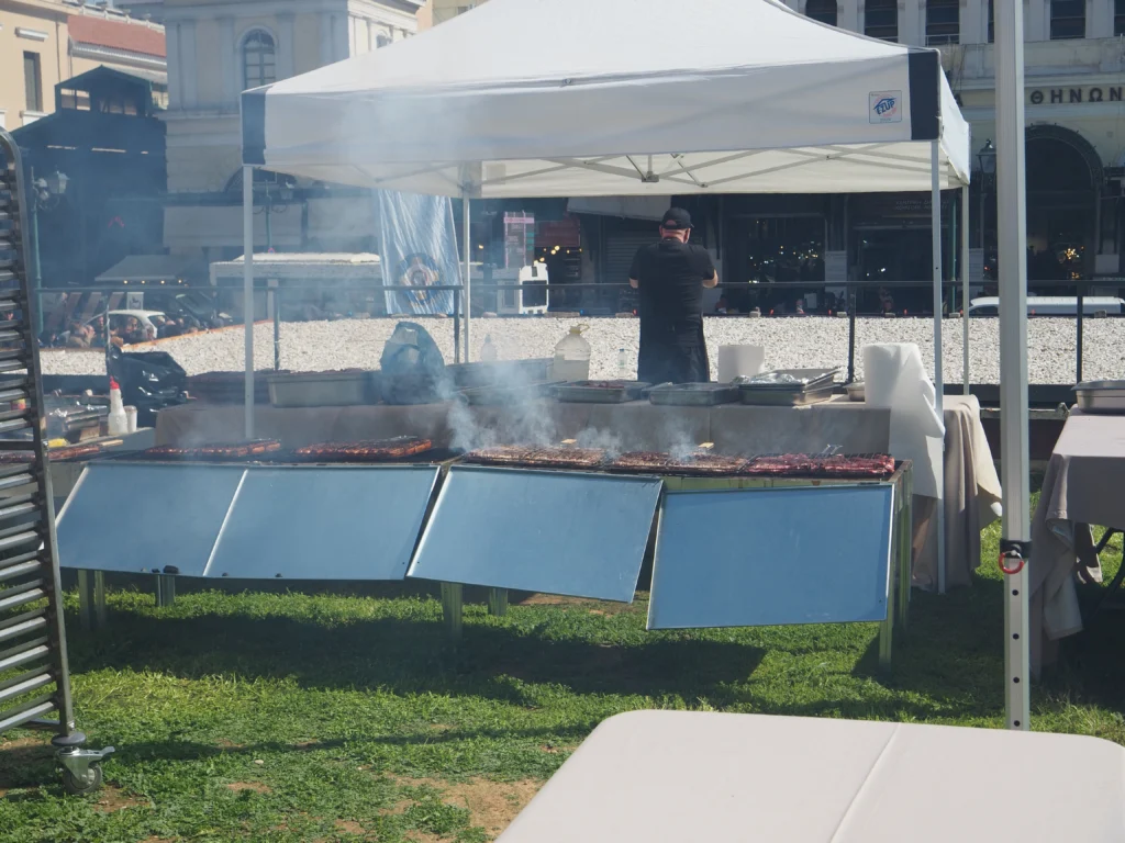 Σχάρες με καλαμάκια που καπνίζουν κάτω από λευκή τέντα σε πλατεία της Αθήνας, με ψήστη γυρισμένο πλάτη