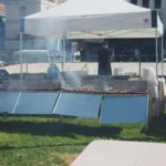 Σχάρες με καλαμάκια που καπνίζουν κάτω από λευκή τέντα σε πλατεία της Αθήνας, με ψήστη γυρισμένο πλάτη