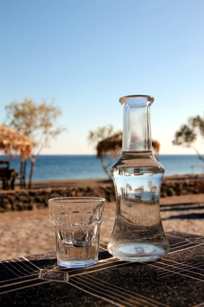 Γυάλινη καράφα και ποτηράκι με τσίπουρο πάνω σε τραπέζι στην παραλία, με θάλασσα και ουρανό στο βάθος