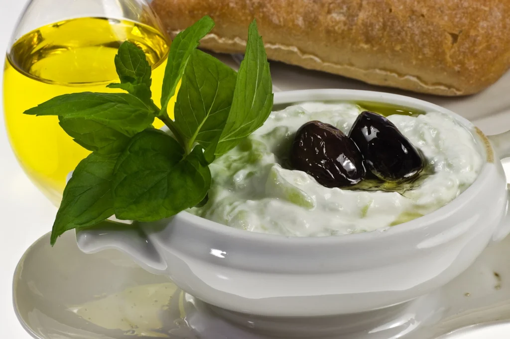 Λευκό μπολ με τζατζίκι, δύο μαύρες ελιές και φύλλα δυόσμου, με ελαιόλαδο και ψωμί πίσω του