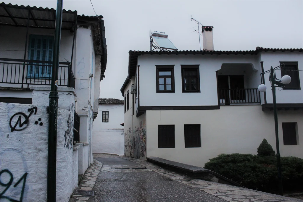 Λιθόστρωτο σοκάκι με λευκά σπίτια και ξύλινα μπαλκόνια στο Βαρούσι, την παλιά πόλη των Τρικάλων, σε συννεφιά