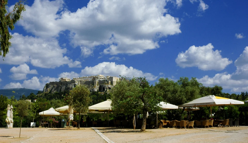 Υπαίθριο καφέ με λευκές ομπρέλες και δέντρα, με τον λόφο της Ακρόπολης στο βάθος